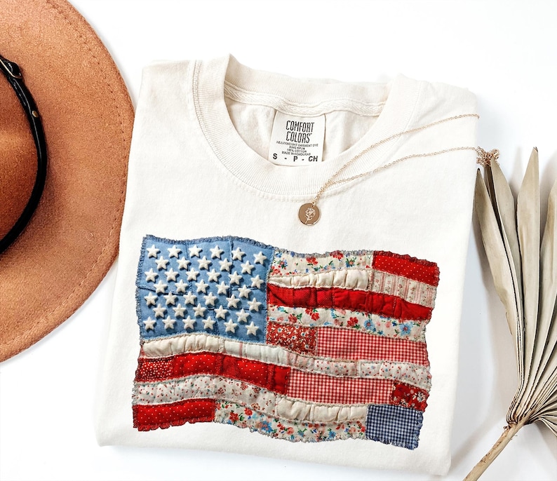 Op de afbeelding: Cr&egrave;mekleurig T-shirt met een patchwork ontwerp van de Amerikaanse vlag. De vlag bevat blauwe denim met witte sterren en rood- en bloempatroon strepen. Een gouden ketting en een bruine hoed zijn ook zichtbaar.