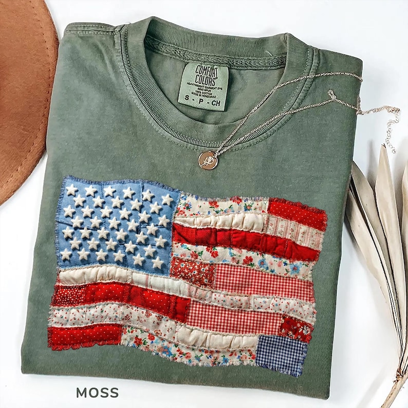 Op de afbeelding: Een mosgroen t-shirt met een opgenaaid Amerikaans vlagontwerp. De vlag heeft een blauw veld met witte sterren en rood-wit gestreepte secties van bedrukte stof. Een zilveren ketting met een hanger ligt op het shirt.