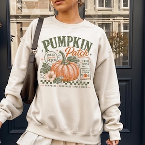 May include: A cream-colored crewneck sweatshirt featuring a graphic print of an orange pumpkin with green leaves. The text reads PUMPKIN Patch, FRESH FROM THE PATCH, FARM FRESH, LOCALLY GROWN, AUTUMN PICKED, PUMPKINS, CORN MAZE, and APPLE CIDER.
