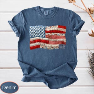 Op de afbeelding: Denim blauw T-shirt met een patchwork Amerikaans vlagontwerp. De vlag bevat witte sterren op een blauw veld en rood-wit gestreepte secties gemaakt van verschillende stoffen met patronen. Het shirt heeft opgerolde mouwen.