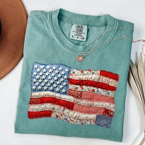 Op de afbeelding: Een zeegroen t-shirt met een patchwork ontwerp van de Amerikaanse vlag. De vlag bevat rode, witte en blauwe stoffen, met een met sterren bezaaid blauw veld en rood-witte strepen. Een zilveren ketting met een hanger ligt op het shirt.