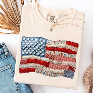 Op de afbeelding: Cr&egrave;mekleurig Comfort Colors T-shirt met een patchwork Amerikaans vlagontwerp. De vlag bevat blauwe denim met witte sterren en rood-wit patronen stoffen strepen. Een zilveren ketting met een hanger ligt op het shirt.