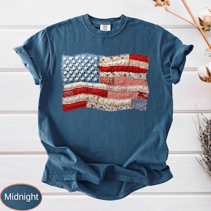 Op de afbeelding: Nachtblau T-shirt met een patchwork Amerikaans vlagontwerp. De vlag gebruikt rood, wit en blauw bedrukte stoffen, waaronder bloemen- en ruitjesprints, met witte sterren op een blauw veld. Het shirt heeft korte mouwen.