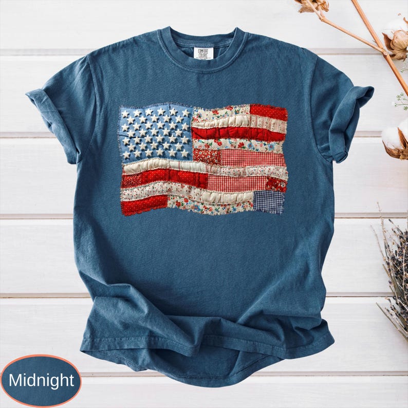 Op de afbeelding: Nachtblau T-shirt met een patchwork Amerikaans vlagontwerp. De vlag gebruikt rood, wit en blauw bedrukte stoffen, waaronder bloemen- en ruitjesprints, met witte sterren op een blauw veld. Het shirt heeft korte mouwen.