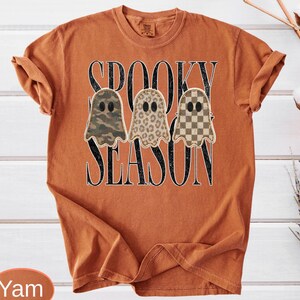 May include: A burnt orange short-sleeve t-shirt featuring the black text SPOOKY SEASON. The design includes three ghost shapes with different patterns: one with green camouflage, one with a brown leopard print, and one with a brown checkerboard.