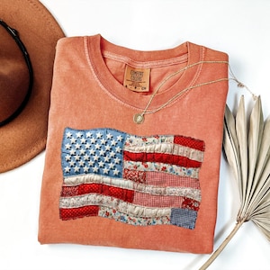 Op de afbeelding: Een koraalkleurig T-shirt met een opgenaaid ontwerp van de Amerikaanse vlag. De vlag heeft een blauw veld met witte sterren en rood-wit gestreepte secties van patroonstof. Een gouden ketting en een bruine hoed staan ook op de foto.