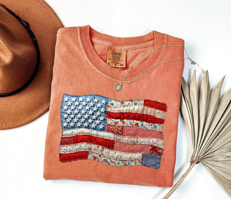Op de afbeelding: Een koraalkleurig T-shirt met een opgenaaid ontwerp van de Amerikaanse vlag. De vlag heeft een blauw veld met witte sterren en rood-wit gestreepte secties van patroonstof. Een gouden ketting en een bruine hoed staan ook op de foto.