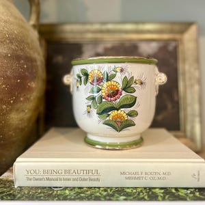 May include: A white ceramic planter with two small handles, featuring a hand-painted floral design in shades of yellow, orange, and green. The rim and base are accented with a green band and a thin gold trim, creating a decorative vessel.