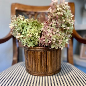 May include: A cylindrical brown bamboo planter with vertical slats holds a large bouquet of dried hydrangea flowers. The flowers display a mix of pale green and soft pink hues, with dense clusters of small petals filling the container.