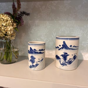May include: Two cylindrical ceramic vessels featuring a hand-painted blue landscape design on a cream-colored background. The artwork depicts stylized mountains, trees, and small structures, with thin blue horizontal bands encircling the top and bottom.