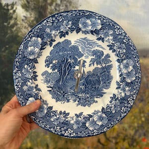 Op de afbeelding: Een blauw-wit bord met bloemenpatroon met een landschapscène in het midden. Het bord heeft een zilveren metalen handvat. Het bord wordt door een hand vastgehouden.