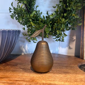 May include: A bronze pear-shaped decorative object with a stem and leaf. The pear has horizontal ridges and sits on a wooden surface. A green wreath and a blue bowl are in the background.
