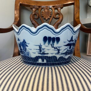 May include: A white ceramic bowl with a scalloped rim, featuring a blue transferware pattern of a landscape with a bridge, a sailboat, willow trees, and a pagoda. The design wraps around the exterior of the bowl in a continuous scene.