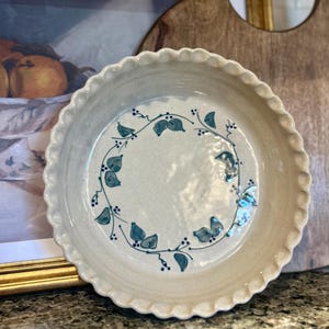 May include: A ceramic pie dish with a scalloped edge. The off-white dish features a blue leaf and berry design around the center. The dish is round and appears to be handmade. A wooden cutting board is in the background.