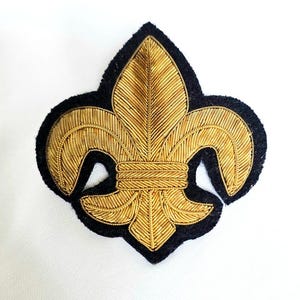 May include: A fleur-de-lis patch featuring intricate gold bullion wire embroidery on a black felt backing. The design showcases a central vertical petal flanked by two curved side petals, all detailed with fine metallic thread textures.