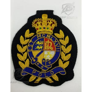 May include: A black embroidered patch featuring a gold crown at the top, a central circular crest with blue, red, and yellow sections, and gold laurel leaves on the sides. The text reads EST MCMLXVII and POLO in gold lettering on blue ribbons.