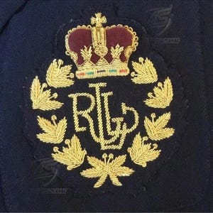 May include: A dark navy blue fabric patch featuring gold embroidered thread detailing. The design includes a central crown with a deep red velvet-like center, the letters RL, and a surrounding wreath of golden leaves stitched onto the fabric.