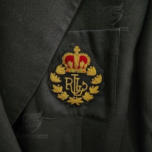 May include: A black blazer featuring a breast pocket with an embroidered crest. The crest displays a red and gold crown above the gold-stitched letters RL, all encircled by a wreath of gold leaves. The fabric has a smooth, dark texture.
