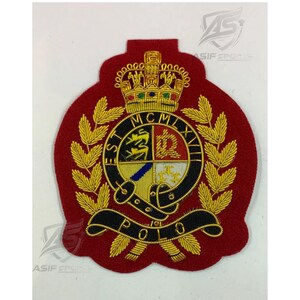 Pode incluir: Um emblema bordado vermelho com uma coroa dourada sobre um brasão circular com o texto EST MCMLXVII e POLO. O brasão inclui um escudo dividido em quatro secções com símbolos, rodeado por folhas de louro douradas e um cinto preto.