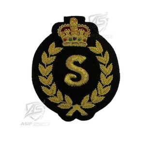 May include: A circular black embroidered patch featuring a gold thread letter S in the center, surrounded by a gold laurel wreath. Above the wreath is a detailed gold crown with red and green accents. The patch has a dark, textured fabric base.