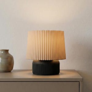 以下が含まれることがあります： ベージュのプリーツシェードと黒いリブベースを備えたモダンなテーブルランプ。 ランプはニュートラルな色の表面にあり、左側に小さな花瓶が見えます。 ランプは柔らかく暖かい光を提供します。