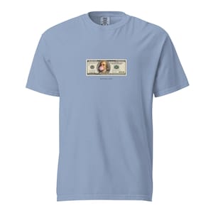 Puede incluir: Una camiseta azul claro con un gráfico de un billete de 100 dólares. La imagen tiene una burbuja de chicle sobre la cara de Benjamin Franklin. La palabra "NOIRLINE" está impresa debajo.