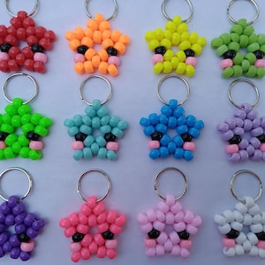 Op de afbeelding: Een verzameling stervormige sleutelhangers in verschillende kleuren, waaronder rood, oranje, geel, groen, blauw, paars, roze en wit. Elke sleutelhanger heeft een kralenontwerp met zwarte en roze accenten en een zilveren sleutelring.