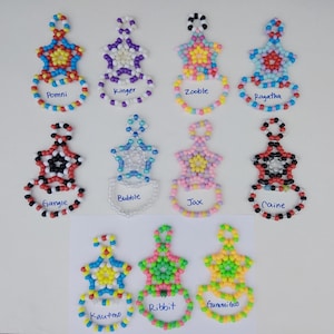 May include: A selection of beaded star-shaped keyrings in various colours. Each keyring has a star design with a ring base, and a name written on the ring. The keyrings are in shades of red, blue, purple, and yellow.