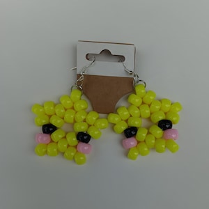 May include: A pair of star-shaped earrings crafted from bright yellow plastic pony beads. Each star features two black beads for eyes and two light pink beads for cheeks, attached to silver-toned metal earring hooks on a brown cardboard display card.