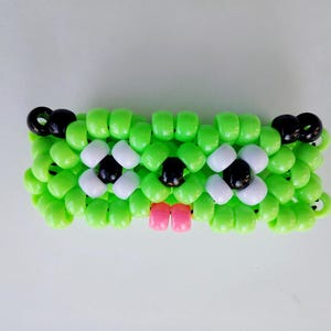 May include: A handmade beaded bracelet in the shape of a frog. The bracelet is primarily lime green with white and black accents for the eyes and pink for the mouth. The bracelet is made of small, round beads.