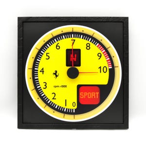 May include: A yellow and black Ferrari-themed clock with a speedometer design. The clock face features a black needle, a red "SPORT" button, and the Ferrari logo. The clock is encased in a black square frame.
