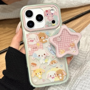 Könnte beinhalten: Transparente Handyhülle mit pastellrosa und mintgrünem Rand. Die Hülle zeigt niedliche Cartoon-Hasen und einen sternförmigen rosa Handyhalter. Der Text "Sug Bunnies" und "cute" sind sichtbar.