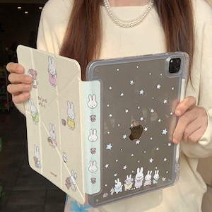 May include: A clear iPad case with a grey background featuring white stars and cartoon bunnies. The case is held open, revealing a beige cover with bunny illustrations. The text "kawaiiXCases" is visible.