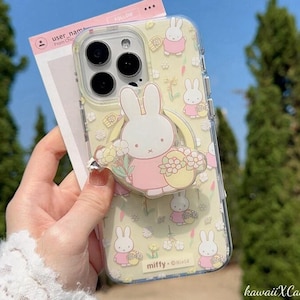 Puede incluir: Funda transparente para teléfono con fondo amarillo y un patrón repetido de conejitos blancos con vestidos rosas. Un agarre para teléfono de conejito blanco con una cesta de flores está adjunto. La funda tiene el texto "miffy - Official".