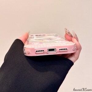 May include: A clear phone case with a pink border and a cartoon design. The case has a transparent back with a pink and white checkered pattern. The case is held in a hand.