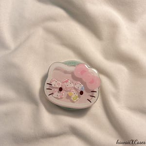 May include: A pink and white phone grip featuring a Hello Kitty design with a pink bow and small character accents. The grip has a clear, circular base and is set against a white fabric background. The text "kawaiiXCases" is in the bottom right corner.