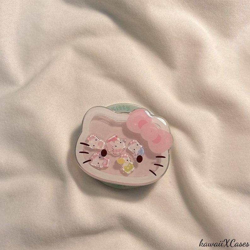May include: A pink and white phone grip featuring a Hello Kitty design with a pink bow and small character accents. The grip has a clear, circular base and is set against a white fabric background. The text "kawaiiXCases" is in the bottom right corner.