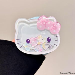 May include: Clear, Hello Kitty-shaped phone case with a pink bow and various character designs inside. The case has a liquid-filled compartment with floating glitter and character figures. The text "kawaiiXCases" is in the bottom right corner.
