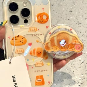 May include: A clear phone case with a cartoon bakery theme, featuring illustrations of a pretzel, croissant, cookie, and coffee. A matching pop socket with a croissant design is attached. The case has the text "INS HOUSE" on a tag.