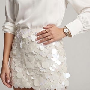 May include: A white long-sleeved blouse paired with a white mini skirt covered in overlapping circular white sequins. The outfit is accessorised with two gold rings on the fingers and a gold-toned wristwatch with a metallic link band.