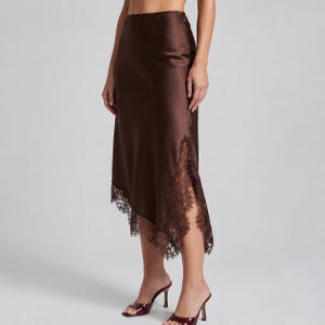 May include: A dark brown midi skirt with a smooth, satin-like finish. The skirt features an asymmetrical hemline adorned with intricate, matching dark brown lace trim that extends along the side and bottom edge of the garment.