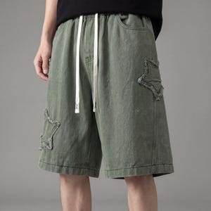 May include: Olive green denim shorts with a drawstring waist and two star-shaped patches. The shorts have a relaxed fit and a white drawstring. The shorts are knee-length.