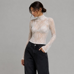 Puede incluir: Una blusa de encaje blanco de manga larga, semi-transparente, con cuello alto y corte ajustado. La prenda está confeccionada en un delicado tejido de encaje floral y se combina con unos pantalones vaqueros de mezclilla en color gris oscuro.