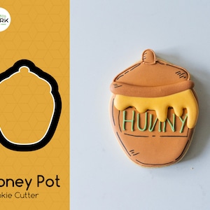 May include: A brown honey pot cookie cutter with a yellow honey drip and the word "Hunny" written on it. The cookie cutter is on a white background.