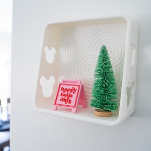 May include: A white shelf with a Mickey Mouse design features a small pink sign that says "happy holla days" and a small green artificial Christmas tree.