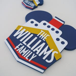 Puede incluir: Un letrero de nombre de familia con tem&aacute;tica de crucero azul y amarillo con el texto "The Williams Family" en letras blancas. El letrero tiene forma de orejas de Mickey Mouse.