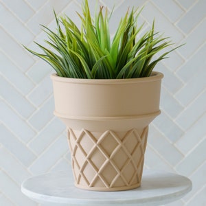 May include: A light brown ceramic planter shaped like an ice cream cone with a green succulent plant inside. The planter is sitting on a white marble surface.