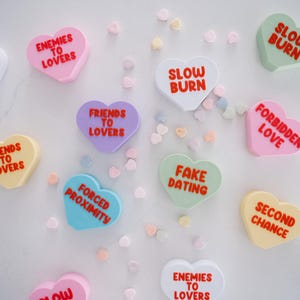 May include: A collection of heart-shaped candy conversation hearts with various messages, including "Enemies to Lovers", "Friends to Lovers", "Slow Burn", "Fake Dating", "Forbidden Love", "Second Chance", and "Forced Proximity".