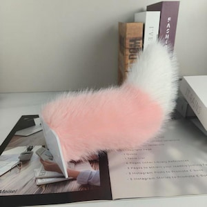 May include: A fluffy, pink and white faux fur tail, likely for cosplay or costume use. The tail is mostly pink with a white tip. The tail is resting on a magazine with text and other books in the background.