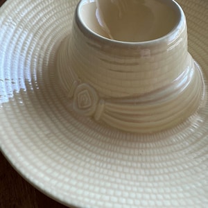 May include: A cream-colored ceramic serving dish with a hat-shaped design. The dish features a wide, textured brim and a central, hat-shaped bowl. A decorative rose detail adorns the hat's band. The dish is designed for serving dips or snacks.